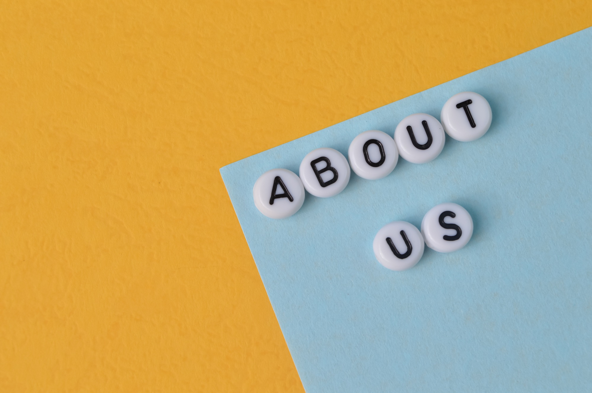 Flat lay of white letter beads spelling 'About Us' on blue and yellow background – representing dental practice storytelling and team introduction